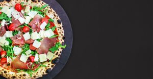 Une pizza alléchante, fraîchement sortie du four à bois, trône sur une assiette en ardoise. Sa pâte croustillante est garnie de jambon italien, de tomates cerises, de roquette et de parmesan râpé, offrant un contraste de couleurs et de textures appétissant. Le fond noir met en valeur la composition harmonieuse de la pizza et son aspect rustique authentique.