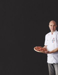 Grégory Edel, champion de France et vice-champion du monde de pizza, présente fièrement sa pizza au jambon italien. La pizza, garnie généreusement de jambon italien de qualité, de mozzarella fondante et de basilic frais, est cuite à la perfection dans un four à bois traditionnel.